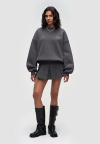 Übergroßer grauer Pullover mit rundem Ausschnitt, kombiniert mit einem grauen, plissierten Minirock und kniehohen schwarzen Stiefeln mit Schnallen-Detail.