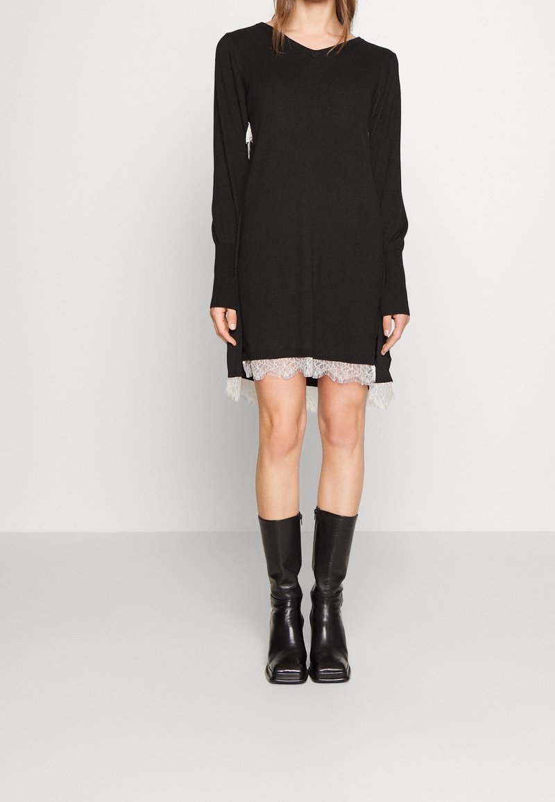 Femme portant une robe noire à manches longues avec un bord en dentelle blanche et des bottes en cuir noires mi-mollet, debout devant un fond clair uni.