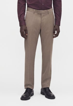 Homme portant un pantalon de costume beige associé à des chaussures habillées noires et une chemise à manches longues bordeaux foncé, debout devant un fond blanc.