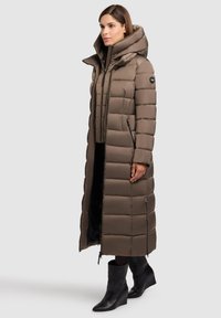 Ilgas rudų pūkuotų paltas su užtrauktuku ir gobtuvu, pasižymintis siuvinėtu dizainu ir šoninėmis kišenėmis, derinamas su juodais kulkšninių batais.