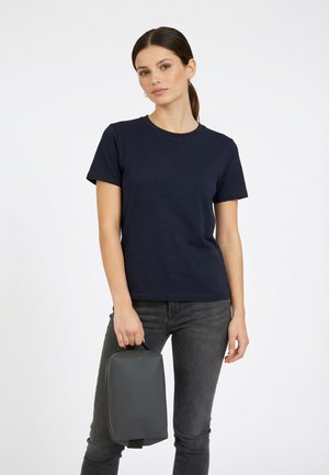 Young woman with dark hair in a ponytail wears a navy t-shirt and gray jeans, holding a dark gray rectangular bag by its handle.