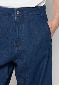 Ciemne spodnie dżinsowe z zapięciem na guziki, bocznymi kieszeniami i prostymi nogawkami. Tkanina ma gładką teksturę z delikatnymi detalami szwów.