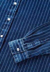 Blaue Jeanshemd mit vertikalen weißen Streifen, ausgestattet mit silbernen Druckknöpfen entlang der Knopfleiste und einem Umschlagärmel mit gestreiftem Muster.