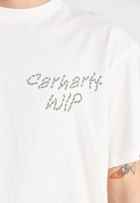 Balta medvilninė marškinėliai su grafikiniu dizainu, kuriame žalios sėklos formos sudaro u�žrašą "Cahert WIP" priekyje. Trumpomis rankovėmis ir apvalia iškirpte.