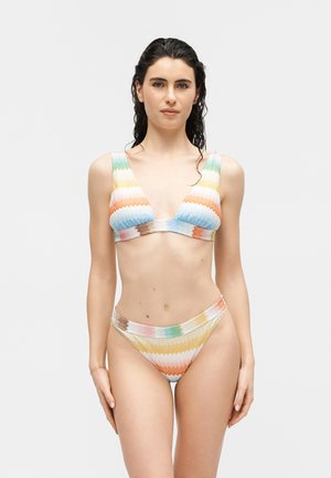 Femeie stând cu fața înainte, purtând un bikini cu model zigzag multicolor, păr ud și închis la culoare, pe un fundal simplu, deschis la culoare.