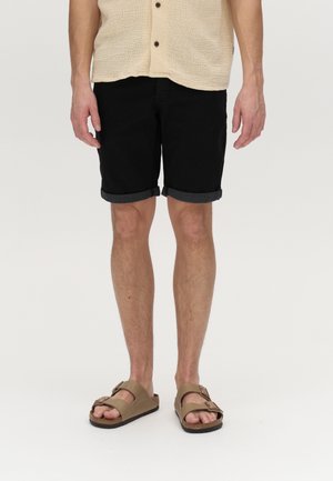 Shorts en denim noir avec ourlets roulés, associés à des sandales marron avec doubles boucles et une sangle texturée.
