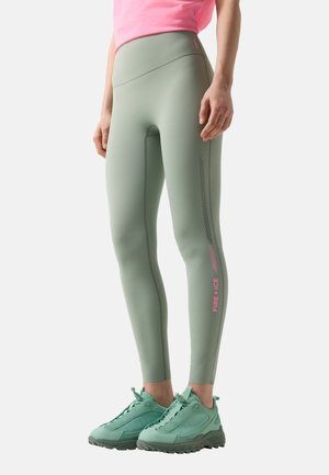 Woman wearing sage green leggings with pink "FIRE + ICE" text on the calf, paired with matching green sneakers and a pink top.