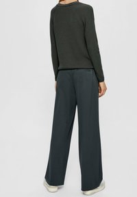 Eine Person trägt einen dunkelgrünen Strickpullover und weite schwarze Hosen, steht mit dem Rücken zur Kamera vor einem schlichten, hellen Hintergrund.