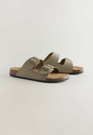 Par sandalov v olivno zeleni barvi, ki se enostavno obujejo, z dvema nastavljivima zaponkama in stopalnim delom iz teksture plute na svetli podlagi.