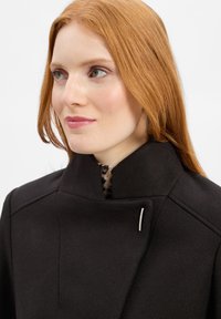 Zwarte wollen jas met hoge kraag, voorzien van een zilveren accentspeld en een patroonvoering die zichtbaar is bij de halslijn. Glad textuur.