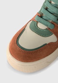 Baskets avec un dessus en daim marron, des accents en cuir vert et une semelle intermédiaire crème. Les caractéristiques incluent des lacets, des détails de couture et une semelle texturée.
