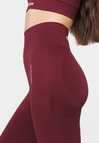 High-waisted burgundy leggings with ribbed texture, mesh accents, and logo detail. Made from stretchy, breathable material.