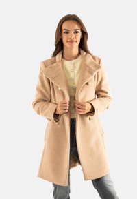 Cappotto in lana beige con un design doppiopetto, grande colletto e bottoni dorati. Indossato sopra un top giallo chiaro e abbinato a jeans grigi.