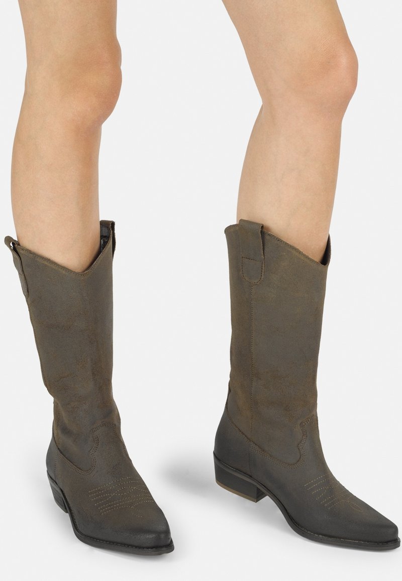 Botas de ante marrones en estilo western con puntas afiladas, un tacón bajo, costuras detalladas y una parte superior con ángulo; cuentan con tirantes laterales para ponerlas fácilmente.