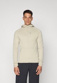 Veste zippée beige clair avec une finition texturée, dotée d'une capuche, de manches longues et d'une poche poitrine. Conçue pour les activités en extérieur.