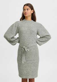 Robe en tricot gris clair, avec un col montant, des manches bouffantes et une ceinture à la taille. Tissu texturé avec des détails côtelés sur l'ensemble.