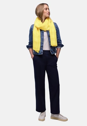 Donna che indossa una sciarpa gialla, giacca di jeans, camicia bianca, pantaloni blu navy e sneakers bianche, in piedi con le mani nelle tasche, che guarda a destra.