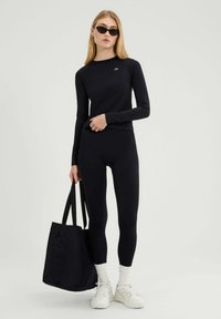 Zwarte lange mouwen top en bijpassende legging in een geribbelde textuur, gecombineerd met een zwarte schoudertas en witte sneakers, tegen een effen achtergrond.