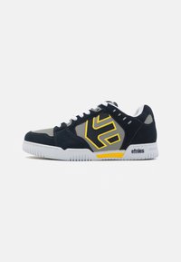 Navy and gray skate shoes with a yellow logo, white laces, rubber soles, and suede accents. Features a low-top design and breathable materials.