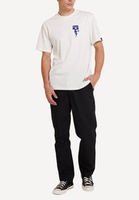 Camiseta de algodón blanca con un gráfico de rayo azul en el lado izquierdo del pecho, combinada con pantalones negros y zapatillas negras con cordones blancos.