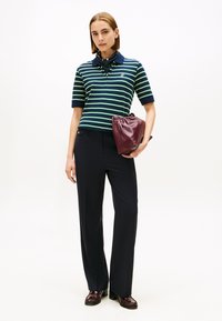 Polo rayé en bleu marine et vert, associé à un pantalon foncé. Le mannequin tient une pochette en cuir bordeaux. Les chaussures présentent un design détaillé.