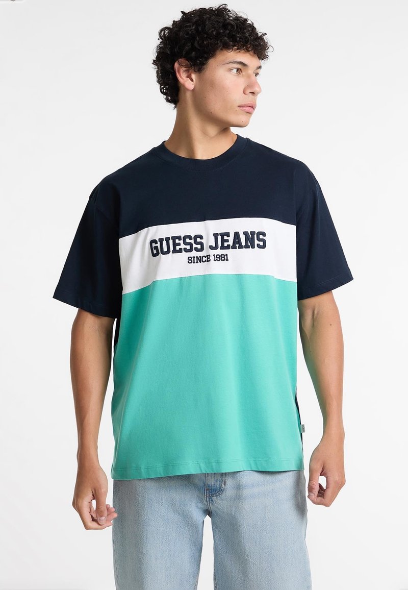 Īsām piedurkņu T-krekls tumšzilā, baltā un teal krāsā. Ar uzrakstu "GUESS JEANS SINCE 1981". Izgatavots no kokvilnas, ar brīvu piegriezumu, sānu šķelumiem un apaļu kakla izgriezumu.