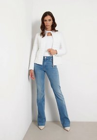 Witte gesteppte jas met een hoge kraag, gecombineerd met een nauwsluitend wit topje en lichtblauwe uitlopende spijkerbroek. Puntige beige schoenen maken de outfit compleet.