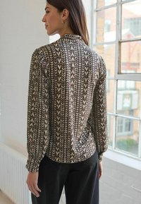 Mønstret bluse med et geometrisk design i creme og brunt, med lange ærmer og en skræddersyet pasform, parret med sorte bukser.