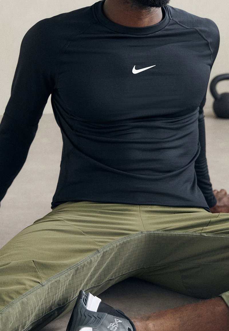 Černé dlouhé rukávy sportovní tričko s bílým logem Nike, v kombinaci se zelenými zúženými kalhotami s texturovaným bočním panelem a černými sportovními botami.