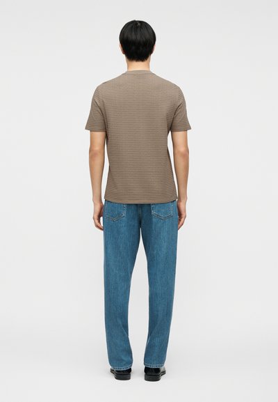 Άνδρας που στέκεται με την πλάτη, φοράει καφέ κοντομάνικο πουκάμισο με υφή, μπλε τζιν και μαύρα παπούτσια μπροστά σε απλό φόντο.