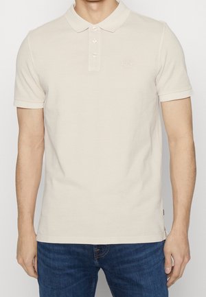 Beżowa bawełniana koszulka polo z trzema guzikami, krótkimi rękawami i subtelnym logo. Połączona z ciemnoniebieskimi dżinsami.