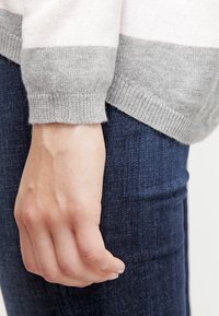 Pull en maille rayé gris et blanc avec une texture douce, doté d'un poignet côtelé, associé à un tissu en denim bleu foncé.