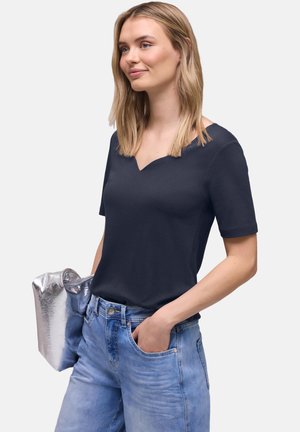 Donna con una maglietta nera a maniche corte e jeans azzurri, che tiene una borsa a mano argento mentre ha una mano in tasca.