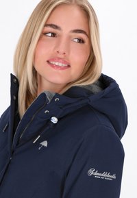 Ženska jakna tamnoplave boje s kapuljačom, siva obloga, srebrni dodaci i logo na rukavu. Glatka tekstura i krojeni dizajn.
