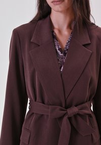 Bruine blazer met ceintuur en brede revers, gemaakt van een gladde stof. Heeft een enkele voorkeurszak en een contrasterende, gedessineerde voering.
