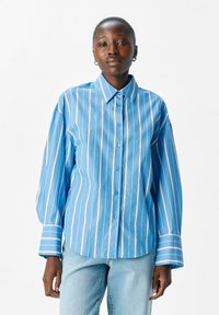 Blau gestreiftes Hemd mit breiten Ärmeln. Verfügbar mit dünnen wei�ßen vertikalen Streifen und einem klassischen Kragen. Kombiniert mit hellblauen Jeans.