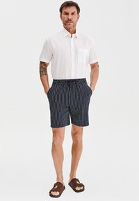 Krótki rękaw, biały lniany koszula z kieszenią na piersi, w połączeniu z granatowymi szortami w paski i brązowymi sandałami. Szorty mają elastyczny pas.