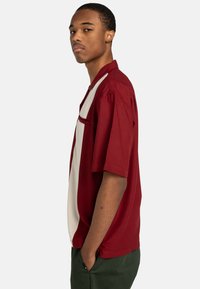 Camicia a maniche corte di un profondo colore rosso con un design a pannello bianco. Presenta un colletto e una tasca sul petto, realizzata in tessuto leggero.
