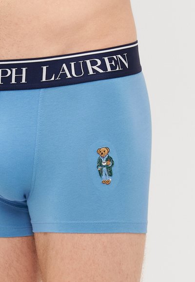 Boxeri din bumbac de culoare bleu deschis, cu un bandou de culoare bleumarin pe care este inscripționat "Ralph Lauren" și un urs brodat în halat pe față.