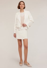Blazer bianco su misura con maniche lunghe, top a strati in tonalità blush e una gonna corta. Tacchi slingback beige a punta completano l'outfit.