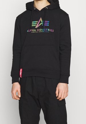 Person, die einen schwarzen Alpha Industries Hoodie mit Regenbogenlogo und Schrift trägt, kombiniert mit schwarzen Cargo-Hosen, vor weißem Hintergrund stehend.