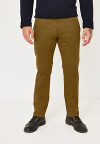 Man wearing mustard-colored pants, black lace-up shoes, and a dark navy sweater standing against a plain background.