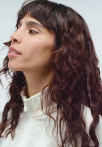 Kučeravé, po plecia dlhé tmavé vlasy, oblečený v bielej bunde s vysokým golierom. Hladká textura, jemný lesk a minimalistický dizajn.