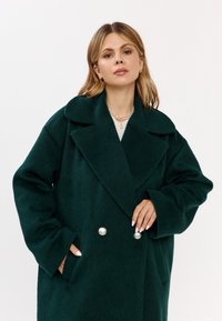 Zielony płaszcz z wełny z kołnierzem z klapami, luźnym krojem, dużymi perłowymi guzikami oraz kieszeniami po bokach. Miękka tekstura i swobodny design.