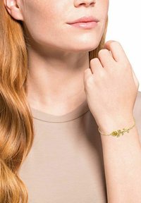 Mujer con cabello largo y rojo que lleva una blusa beige, mostrando una pulsera de oro con una rosa y hojas en su muñeca.