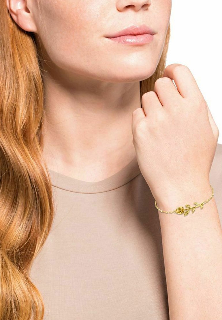 Mujer con cabello largo y rojo que lleva una blusa beige, mostrando una pulsera de oro con una rosa y hojas en su muñeca.