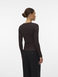 Frau mit braunen Haaren im Dutt trägt ein langärmliges, dunkelbraunes Oberteil und schwarze Hosen, steht mit dem Rücken zu einem weißen Hintergrund.