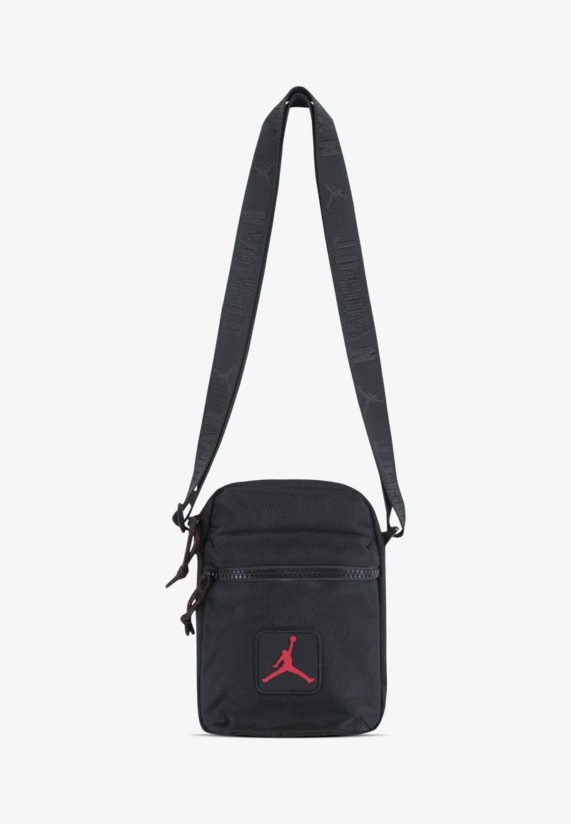 Saco crossbody preto em nylon com alça ajustável, fechos de zíper e um logo vermelho da Jordan na parte frontal. Superfície texturizada, design compacto.