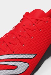 Κοντινό πλάνο ενός λαμπερού κόκκινου αθλητικού παπουτσιού με υφή υφάσματος, κόκκινα κορδόνια και μεγάλο λευκό και μαύρο λογότυπο στο πλάι.