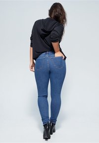 Nainen, joka on pukeutunut sinisiin farkkuihin ja mustaan pitkähihaiseen huppariin, seisoo yksi käsi takataskussa ja mustat nilkkakorkokengät jalassa.